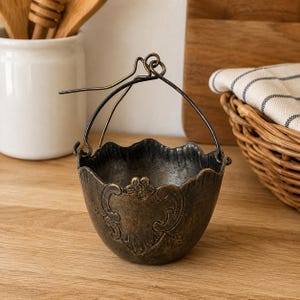 Könnte beinhalten: Eine kleine Schale aus dunkel bronzefarbenem Metall mit gewelltem Rand und einem dekorativen, geprägten Blumenmuster auf der Vorderseite. Die Schale hat einen dünnen, gebogenen Metallgriff, der an zwei kleinen Ösen an den Seiten befestigt ist.