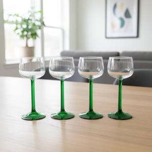 Puede incluir: Cuatro copas de vino transparentes con tallos verdes dispuestas sobre una mesa de madera clara. Las copas tienen forma de cuenco redondeado y base plana. El fondo incluye una ventana, una planta y un sofá gris.