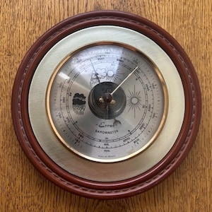 May include: A round barometer with a dark brown wooden frame and a brass-colored inner ring. The face displays weather symbols, a needle, and measurements in inches, millibars, and millimeters. The text "Erref Barometer" is visible.