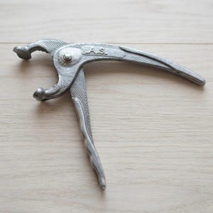 Pode incluir: Um abridor de potes de metal prateado com uma superfície de aderência texturizada nas alças. A ferramenta possui um mecanismo de mandíbula curva para agarrar tampas e tem as letras A.S. gravadas na alça superior perto do pivô.