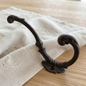 Könnte beinhalten: Ein dunkelbrauner Wandhaken aus Metall mit geschwungenem, schnörkelartigem Design und einer verzierten, strukturierten Oberfläche. Der Haken verfügt über eine breite, flache Basis mit einem sichtbaren Schraubloch zur Montage und schwingt elegant.