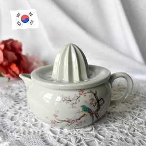 Op de afbeelding: Witte keramische citruspers met een ruimer en een kleine kan. De citruspers is versierd met een kleurrijk vogel- en bloemmotief. De citruspers heeft een handvat en een tuit.