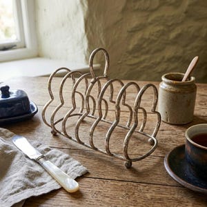 May include: A metal toast rack with a distressed silver finish, featuring six arched slots for holding slices of bread. The rack has a central loop handle at the top and rests on small rounded feet, designed for organising and serving toast at the table.