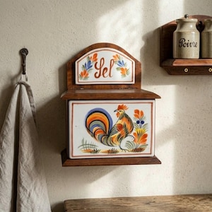 Può includere: Una scatola per il sale in legno da parete con un pannello frontale in ceramica raffigurante un gallo colorato e una placca superiore in ceramica con la scritta Sel. La scatola è dotata di un coperchio incernierato ed è vicina a un panno.
