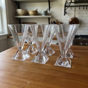 Op de afbeelding: Een set van vijf heldere glazen bekers met een V-vormige kom met verticale ribbels en een gefacetteerde, geometrische vierkante voet. De glazen zijn in een groep gerangschikt, wat het transparante materiaal en het hoekige ontwerp benadrukt.