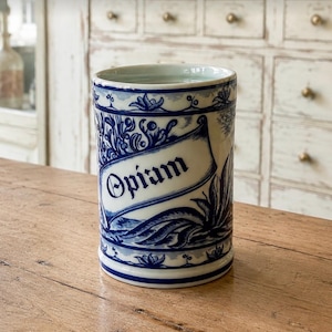 May include: A cylindrical ceramic apothecary jar featuring a white base with intricate blue floral and leaf patterns. A central banner displays the text Opiam in a bold, dark blue gothic-style font, framed by decorative borders at the top and bottom.