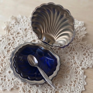 May include: A silver-toned metal clam shell-shaped dish with a hinged lid. The interior of the bottom shell is a deep, glossy cobalt blue. A small, matching silver-toned metal spoon with an etched handle rests inside the blue basin.