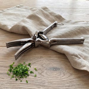 Pode incluir: Um picador de ervas de metal prateado com um design de múltiplas lâminas e um cabo texturizado com padrão de hachura cruzada. A ferramenta é construída com um mecanismo de dobradiça e uma mola, exibida ao lado de ervas verdes picadas.