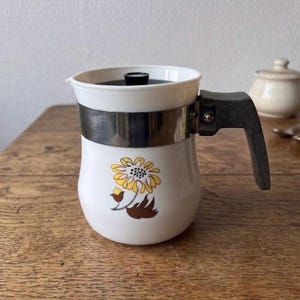 May include: White ceramic coffee pot with a black handle and lid. A silver metal band wraps around the middle. The pot features a yellow and brown floral design. A small sugar bowl is in the background.