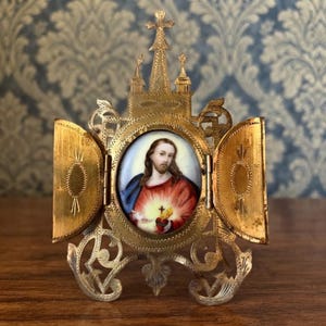 Puede incluir: Un marco tríptico de metal dorado con un retrato ovalado de una figura de cabello castaño largo, que viste una túnica roja y una capa azul, sosteniendo un corazón en llamas con una cruz. El marco tiene detalles recortados y una cruz arriba.