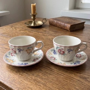 Può includere: Due tazze da tè bianche e piattini con motivi floreali. Le tazze e i piattini hanno un bordo rosa e sono decorate con fiori rossi, blu e gialli. Le tazze da tè sono su un tavolo di legno.