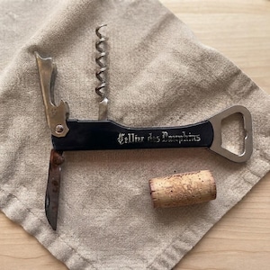 Op de afbeelding: Een zwarte metalen kelnerskurkentrekker met een zilverkleurige spiraal, een flesopener en een klein inklapbaar mesje. Op het handvat staat de tekst Cellier des Dauphins in gotisch lettertype. Een natuurlijke bruine kurk ligt onder het gereedschap.