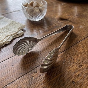 May include: Silver-toned sugar tongs with shell-shaped ends, resting on a wooden surface. A small glass bowl filled with brown sugar cubes is in the background, along with a lace-trimmed cloth. The tongs are a vintage kitchenware item.