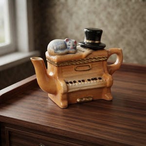 Peut inclure: Une théière en céramique en forme de piano droit avec une finition beige brillante. Un petit chat endormi aux motifs bleus et blancs repose sur le couvercle supérieur à côté d'un haut-de-forme noir avec une bande dorée. La théière a un bec et une anse.