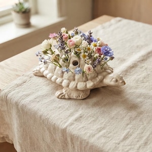 Op de afbeelding: Een crèmekleurige, schildpadvormige keramische bloemenvaas met meerdere ronde openingen in het schild. De vaas is gevuld met een verscheidenheid aan kleine verse bloemen, waaronder roze rozen, witte madeliefjes, blauwe korenbloemen en takjes lavendel.