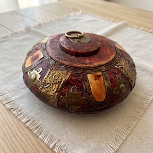 May include: A round, reddish-brown wooden box with a lid featuring a central brass ring handle. The exterior is decorated with alternating brass plates embossed with floral and geometric patterns, interspersed with smooth, light-colored bone inlays.