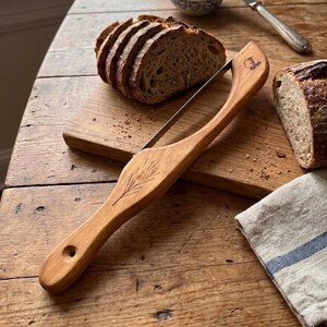 May include: A wooden bread slicer with a stainless steel blade, resting on a wooden cutting board. The slicer has a decorative leaf design. Sliced bread and a loaf of bread are also visible on a wooden table.