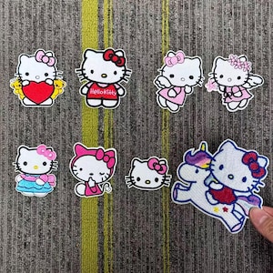 May include: Eight Hello Kitty embroidered patches are displayed. The patches feature the character in different poses, including holding a heart, wearing a dress, and riding a unicorn. Each patch has a white background and a colourful design.