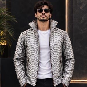 Puede incluir: Una chaqueta plateada y gris con un patrón texturizado similar a escamas en todo el exterior. La chaqueta tiene un cuello alto estructurado, mangas largas y un cierre de cremallera central, usada sobre una camiseta blanca y pantalones oscuros.