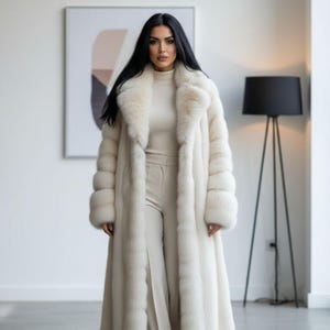 Könnte beinhalten: Ein langer, elfenbeinfarbener Pelzmantel mit breitem Kragen und Manschetten. Der Mantel wird über einem passenden Rollkragenpullover und einer Hose getragen. Das Outfit befindet sich in einem hellen Raum mit einer modernen Stehlampe.