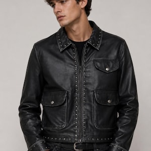 May include: Black leather jacket with silver stud accents along the zipper, collar, cuffs, and pockets. The jacket has two chest pockets and two lower pockets. The model is wearing black jeans and a black shirt.