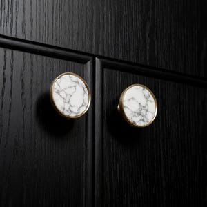 May include: Two round cabinet knobs with white marble faces and gold-colored rims are mounted on a black wooden cabinet door. The marble has gray veining, and the knobs are evenly spaced.