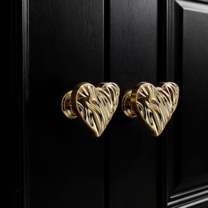 Puede incluir: Primer plano de dos pomos dorados en forma de corazón montados en una puerta de armario negra. Los pomos tienen un diseño texturizado y acanalado y un acabado brillante. La puerta del armario es de madera y tiene una superficie lisa y brillante.