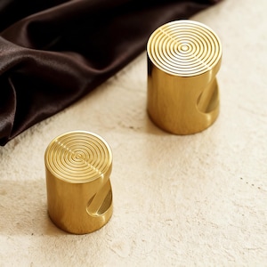 May include: Two cylindrical, gold-colored cabinet knobs. Each knob has a circular, concentric design on the top and a curved cutout on the side for gripping. The knobs are made of a shiny, metallic material.