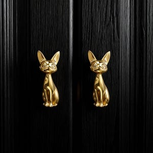 May include: Two gold cat-shaped door handles fixed to a black wooden door. The handles are designed as sitting cats, with pointed ears and smiling faces. The door has vertical wood grain, adding texture to the surface.