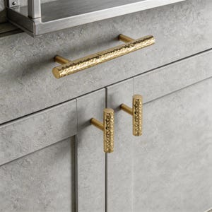 May include: Close-up of gold cabinet hardware. The image shows a long bar handle and two smaller T-shaped pulls. The hardware has a hammered texture and is mounted on a gray cabinet. The hardware is made of metal.