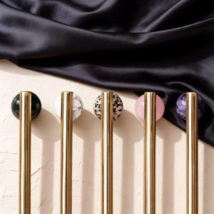 Puede incluir: Cinco tiradores decorativos para armarios con tallos dorados y tapas esféricas en varios colores y patrones. Las tapas incluyen verde, mármol blanco, moteado, rosa y morado. Los tiradores están colocados sobre un fondo neutro con una tela oscura drapeada.