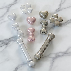 Pode incluir: Uma variedade de puxadores decorativos para armários em várias formas, incluindo corações, trevos e desenhos em forma de osso. Os puxadores são em tons de prata e rosa, com um acabamento brilhante. Os puxadores estão dispostos sobre uma superfície de mármore branco.