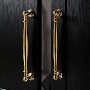 Puede incluir: Dos tiradores dorados de armario con un diseño clásico están montados en una puerta de armario negra. Cada tirador presenta un mango largo y delgado con detalles decorativos y un pomo redondo en cada extremo. Los tiradores son simétricos.