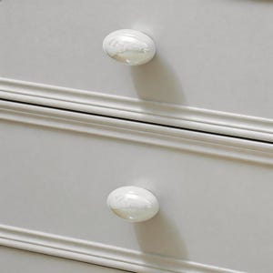 Pode incluir: Close-up de uma cômoda cinza clara com dois puxadores ovais brancos. Os puxadores têm um acabamento perolado e estão centrados nas gavetas. A cômoda tem um design simples e clássico.