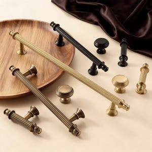 May include: An assortment of cabinet hardware in various styles and finishes. The collection includes long bar pulls, knobs, and smaller pulls in gold, bronze, and black. The hardware is arranged on a wooden tray and a neutral surface.