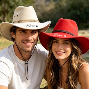 May include: Two cowboy hats, one cream-colored and one red, are shown. The cream hat has the name "Kitty" and a paw print, while the red hat has the name "Tom" and a paw print. Both hats have a brown rope detail and chin straps.