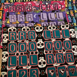 May include: A stack of colorful fabric squares with embroidered letters and numbers. The squares feature various patterns, including skulls, hearts, and lava lamps. The letters spell out "RRR LLL" and "LOL", with numbers 1, 2, and 3.