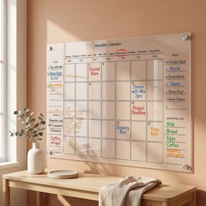 Könnte beinhalten: Ein transparenter, an der Wand montierter Monatskalender aus Acryl mit schwarzen Rasterlinien und gedrucktem Text. Er enthält Bereiche für Aufgaben, Notizen, einen Wochenplan und eine Einkaufsliste mit handschriftlichen Einträgen.