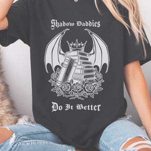 Peut inclure: T-shirt noir avec l'inscription "Shadow Daddies" au-dessus d'un dessin blanc de livres, d'ailes et de roses, avec la phrase "Do It Better" en dessous. La personne porte également un jean bleu.