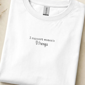 Op de afbeelding: Wit T-shirt met de tekst "I support women's Wrongs" in zwart. Het shirt is opgevouwen en ligt op een neutraal gekleurd oppervlak. Een klein label is zichtbaar op de kraag. Een boek en gedroogde planten zijn op de achtergrond.