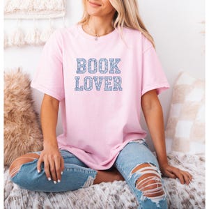 Może przedstawiać: Jasnoróżowa koszulka z napisem "BOOK LOVER" w niebieskich, kwiatowych literach blokowych. Osoba ma na sobie niebieskie dżinsy z podartymi kolanami.