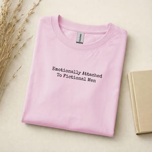 Może przedstawiać: Jasnor&oacute;żowa koszulka Gildan z czarnym napisem "Emotionally Attached To Fictional Men". Koszulka jest złożona na jasnej powierzchni. Widoczna jest gałązka suszonej trawy.