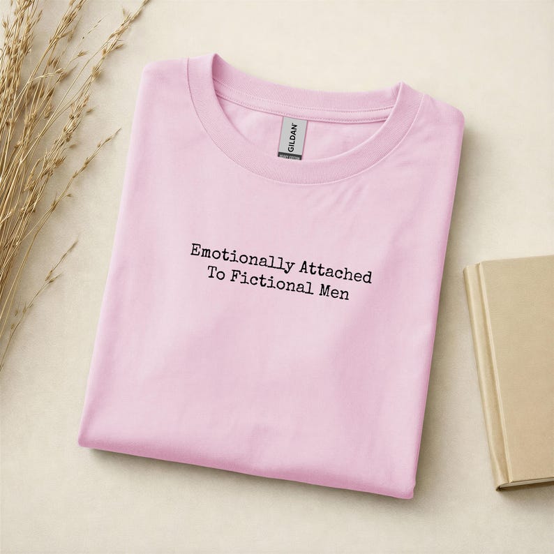 Może przedstawiać: Jasnor&oacute;żowa koszulka Gildan z czarnym napisem "Emotionally Attached To Fictional Men". Koszulka jest złożona na jasnej powierzchni. Widoczna jest gałązka suszonej trawy.