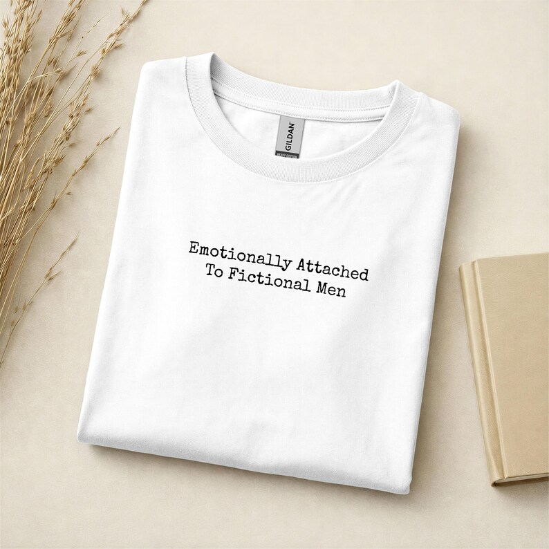 Może przedstawiać: Biała koszulka z czarnym napisem "Emotionally Attached To Fictional Men". Koszulka jest starannie złożona na jasnej powierzchni, z książką i suszonymi roślinami w tle.