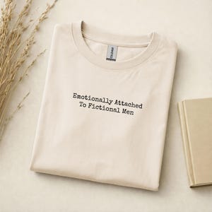 Może przedstawiać: Kremowa koszulka z czarnym napisem "Emotionally Attached To Fictional Men". Koszulka jest starannie złożona. Książka i suszone rośliny znajdują się w tle. Etykieta koszulki jest widoczna na dekolcie.