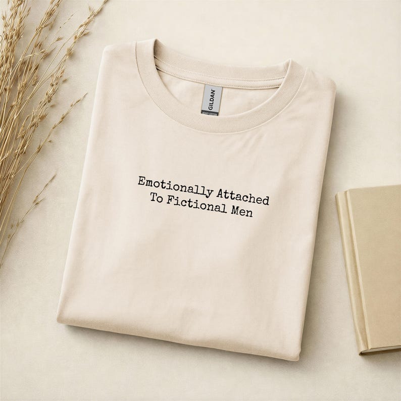 Może przedstawiać: Kremowa koszulka z czarnym napisem "Emotionally Attached To Fictional Men". Koszulka jest starannie złożona. Książka i suszone rośliny znajdują się w tle. Etykieta koszulki jest widoczna na dekolcie.