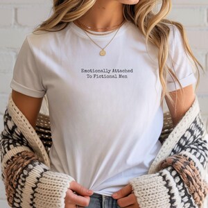 Może przedstawiać: Biały t-shirt z czarnym napisem "Emotionally Attached To Fictional Men". Koszulka jest noszona ze złotym naszyjnikiem i wzorzystym kardiganem.