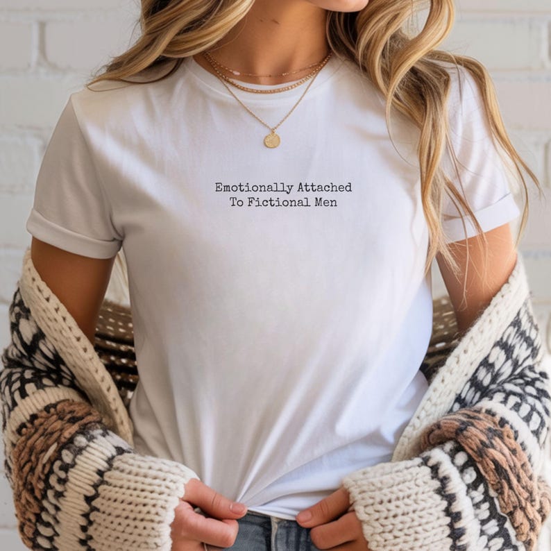 Może przedstawiać: Biały t-shirt z czarnym napisem "Emotionally Attached To Fictional Men". Koszulka jest noszona ze złotym naszyjnikiem i wzorzystym kardiganem.