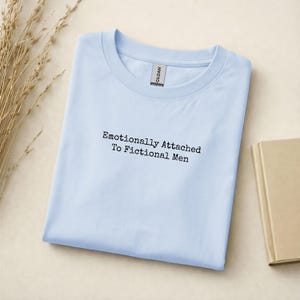Może przedstawiać: Jasnoniebieska koszulka z czarnym napisem "Emotionally Attached To Fictional Men". Koszulka jest starannie złożona na jasnobeżowej powierzchni. Etykieta Gildan jest widoczna przy dekolcie.