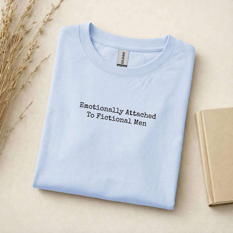 Może przedstawiać: Jasnoniebieska koszulka z czarnym napisem "Emotionally Attached To Fictional Men". Koszulka jest starannie złożona na jasnobeżowej powierzchni. Etykieta Gildan jest widoczna przy dekolcie.